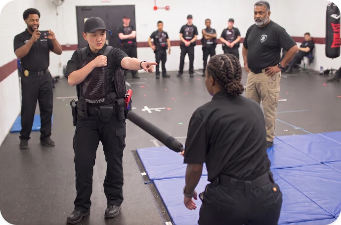 Self-defense training class with an instructor demonstrating a padded baton technique to a participant as others observe.